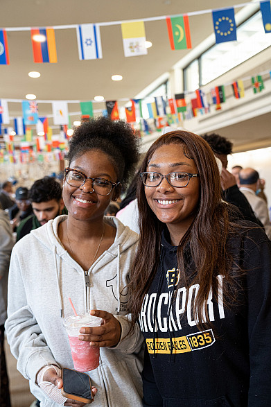 Two students smiling for a photo with flags from different countries in the background