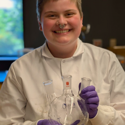 Avery Di Iulio dressed in lab gear in a lab holding a glass beaker and posing for the camera