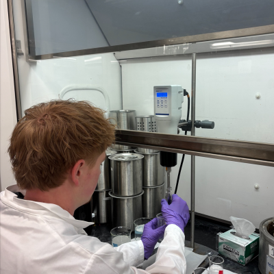 Owen Hazard dressed in lab gear in a lab handling a special instrument that drops liquid into a glass beaker