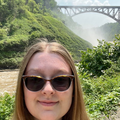 Selfie of Alexis Risdale at a park with a river and bridge in the background