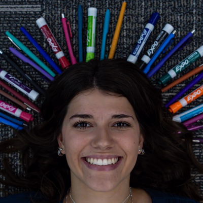 Alanna Pellicane laying on the ground smiling with various markers and pens around her head