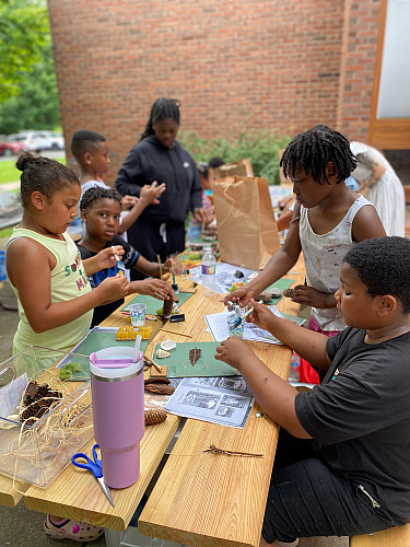 Brockport Summer Learning students making crafts with various outdoor materials at a picnic table outdoors.
