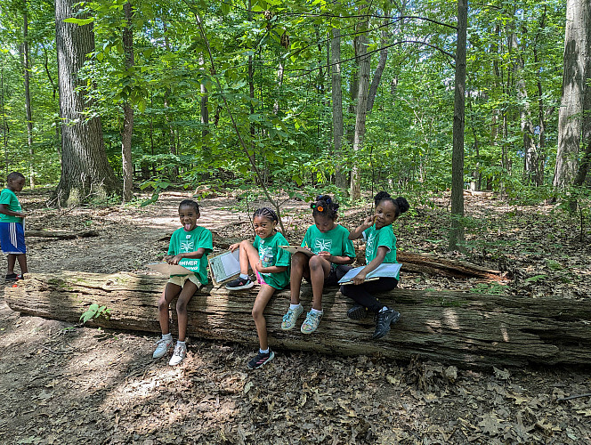 4 Brockport Summer Learning students sitting on a fallen down tree log at the Hamlin Beach State Park.