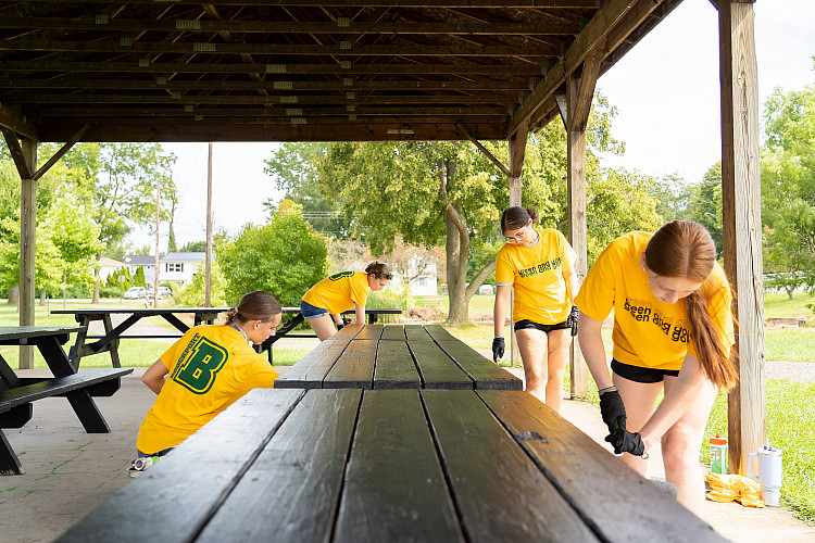 Four students painting picnic tables at a local park