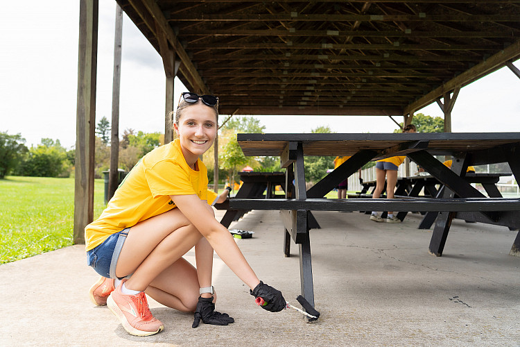 One student, smiling, painting a picnic table at a local park
