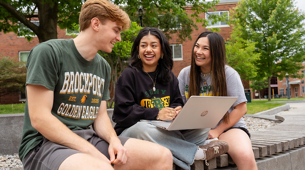 Three Brockport students smiling, looking at laptop.