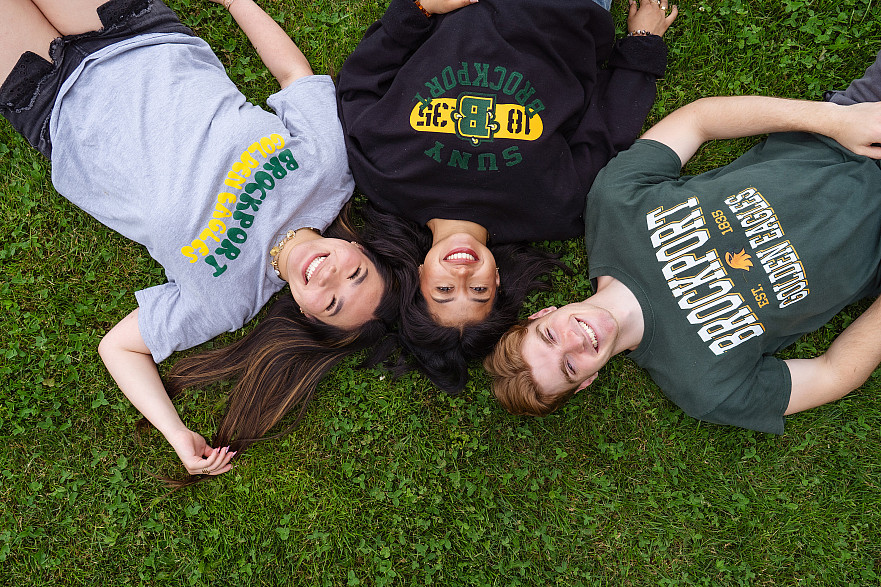 Three Brockport students laying in the grass and smiling at the camera in SUNY Brockport apparel.