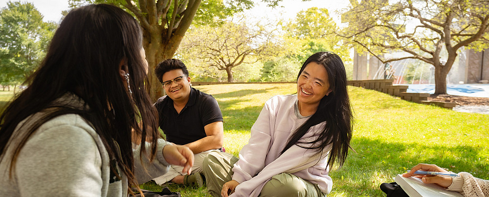 Three SUNY Brockport students laughing together while sitting on the grass outside.