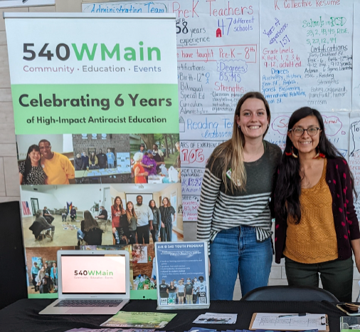Two people standing by table with banner 540WMain Celebrating 6 years of high impact anti-racism education
