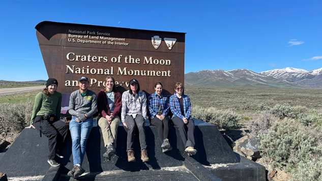 Geology students sitting in front of the Craters of the Moon National Monument sign