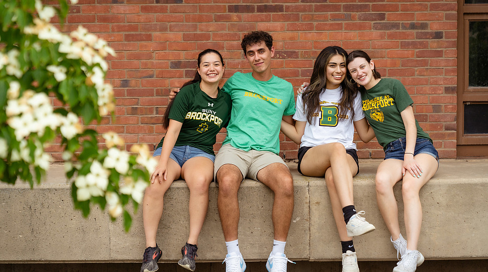 Four students sitting in front of a brick building on the SUNY Brockport campus.