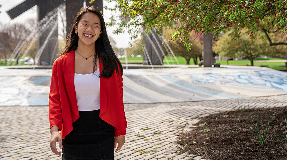 Photo of an Honors Scholar, Lily Wegerski, walking on campus in front of the Special Olympics Fountain in the Spring time