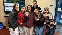 A group of students stand indoors on a campus lobby floor, posing together while holding donuts.