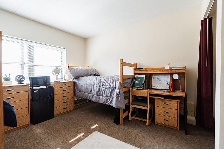 Wide view of a corridor style bedroom with a mini fridge, microwave, dresser, bed, and desk