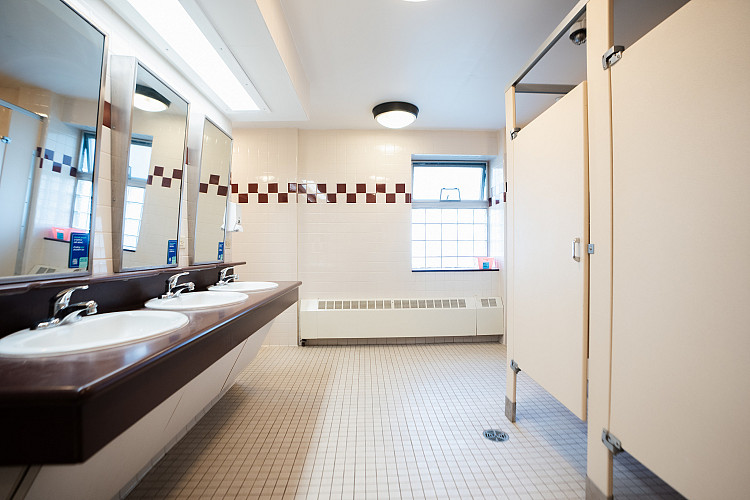Wide view of corridor style bathroom with 3 mirrors and sinks to the left and 2 bathroom stalls to the right