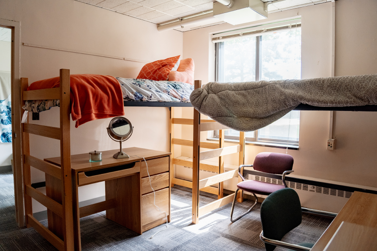 Wide view of a lifted bed with a desk underneath, 2 chairs, and another lifted bed in a corner of a 4 person suite