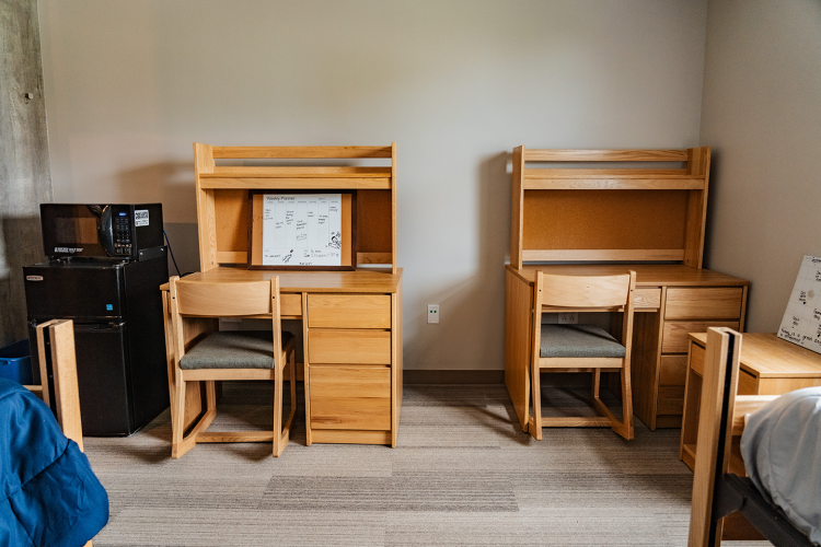 Wide view of a wall away from the beds of a fridge, microwave, and 2 desks with chairs