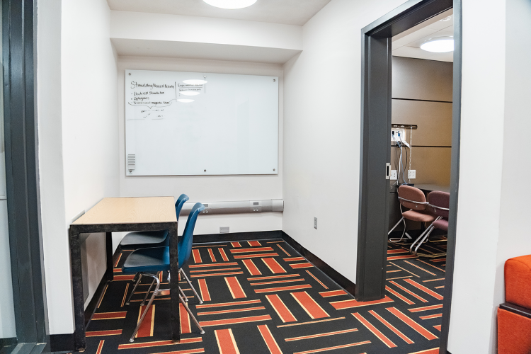Corridor adjacent to a lounge with a table, chair, and a whiteboard on a wall