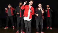 Five singers are dressed in red and black, standing in a v-formation, and are singing into microphones against a dark background.