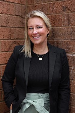 Woman with shoulder length blond hair against a brick wall wearing a black shirt and a black blazer